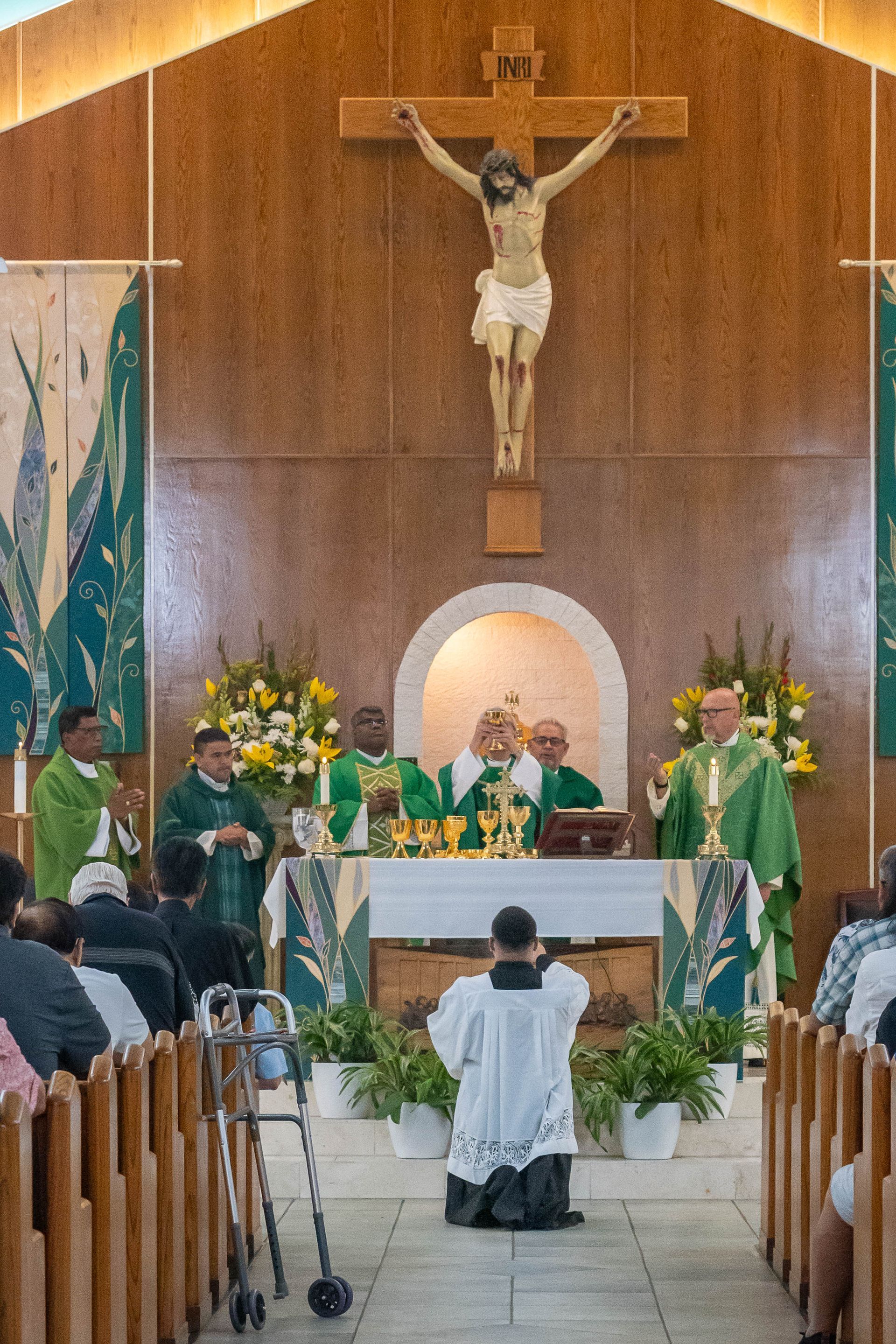 A group of clergy in green vestments stands at a church altar beneath a crucifix, with one person kneeling in the foreground.