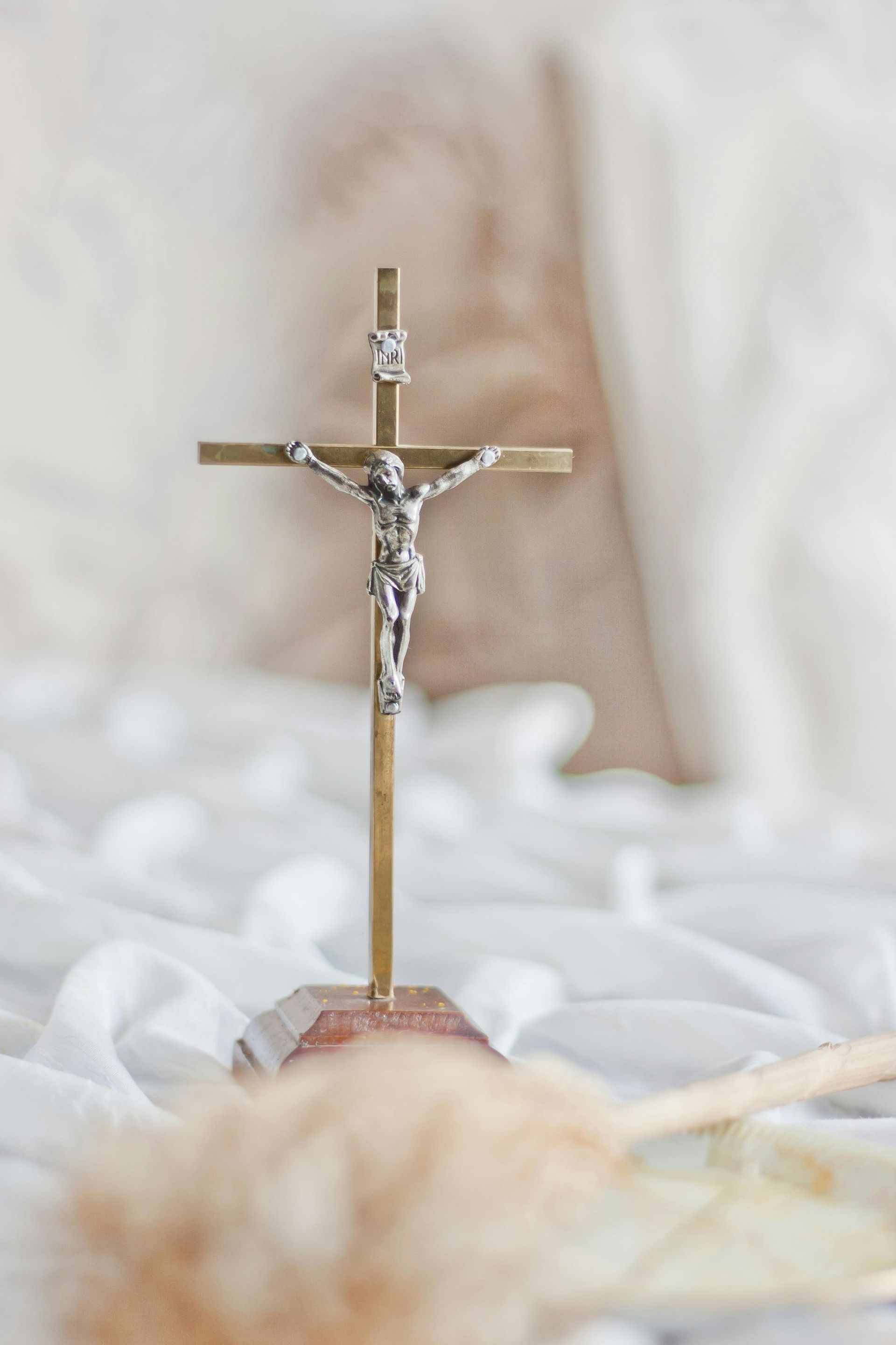 A silver crucifix standing on a base against a soft, white, draped fabric background with blurred decor in the foreground.
