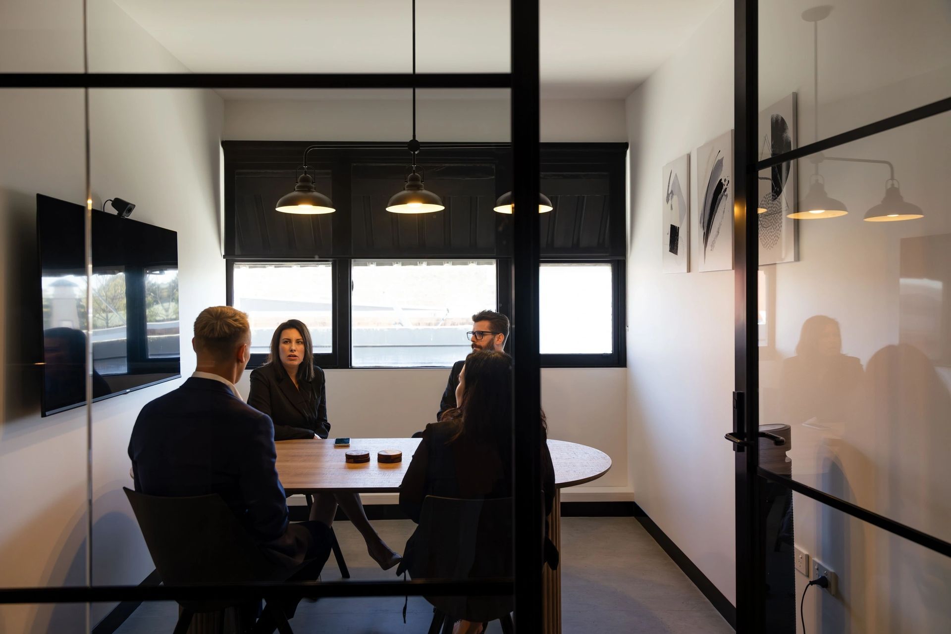 Four people in a meeting around a table in a modern office with glass walls and a TV.
