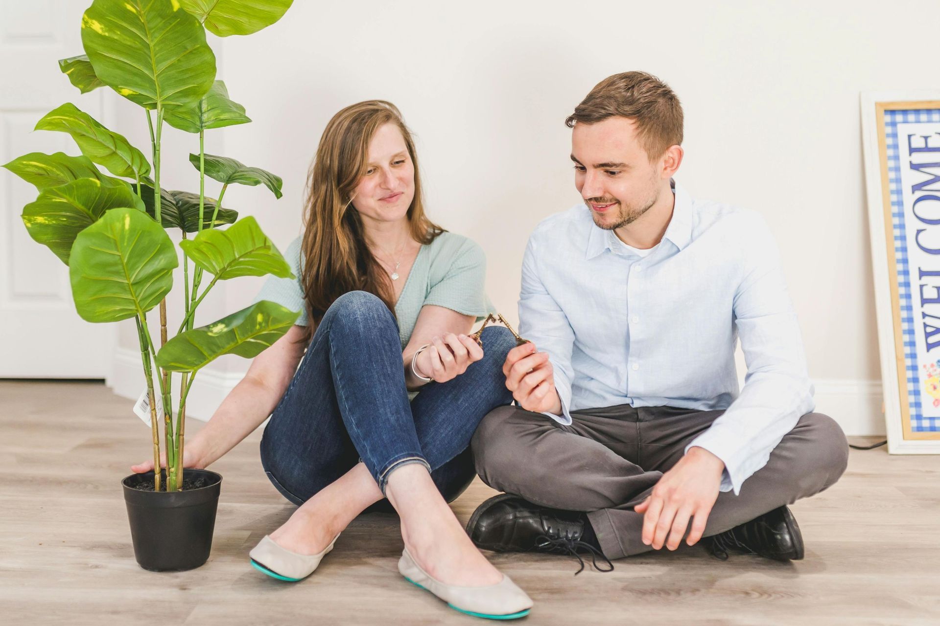 Woman and man seated on floor with plant, smiling, near a welcome sign.