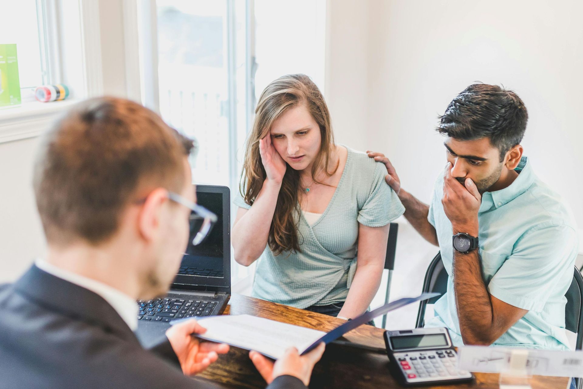 Couple looking concerned at documents with a person in a suit, a calculator, and a laptop.