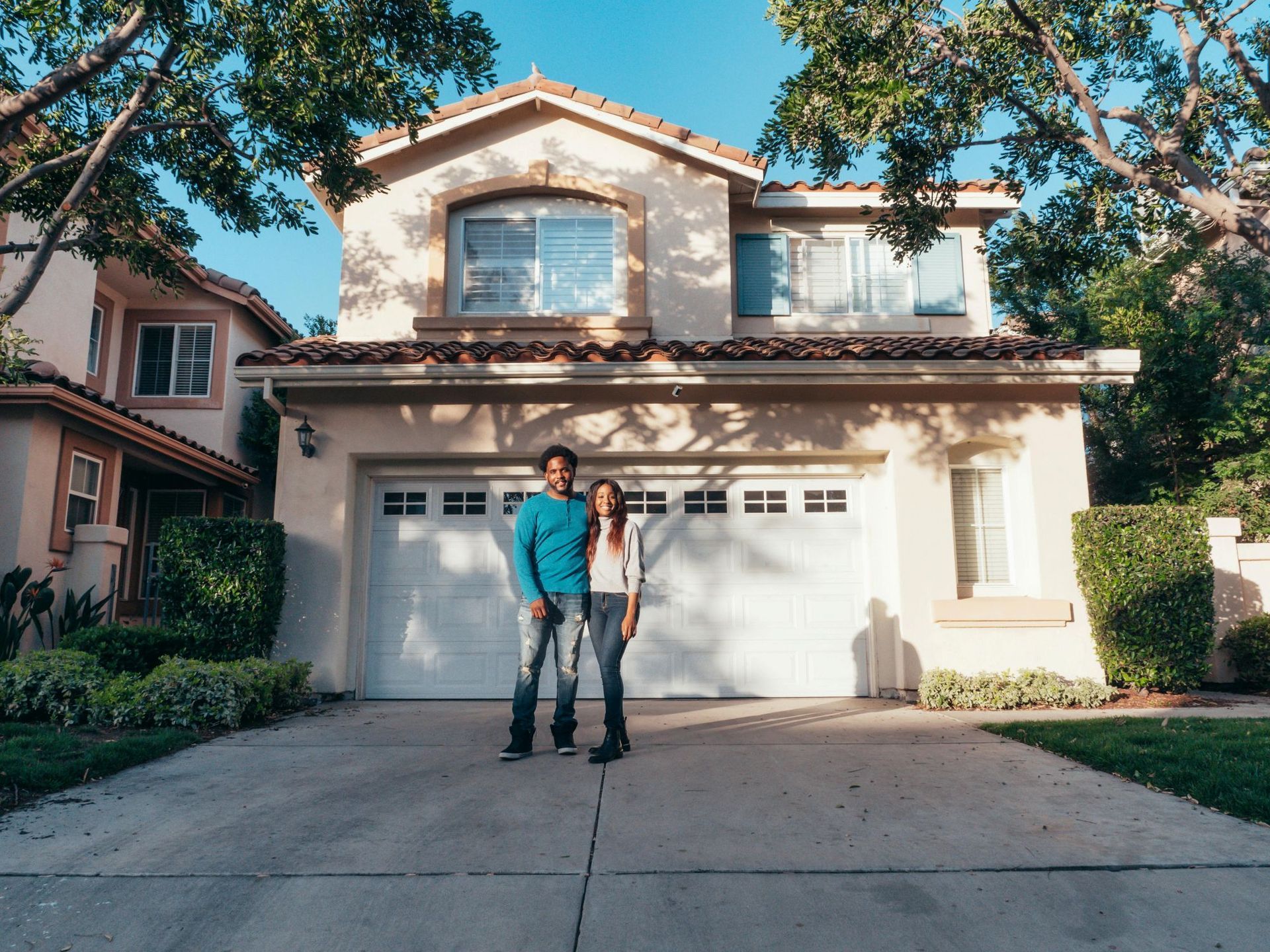 Couple standing in front of a house, driveway, and garage. Bright day, trees around.