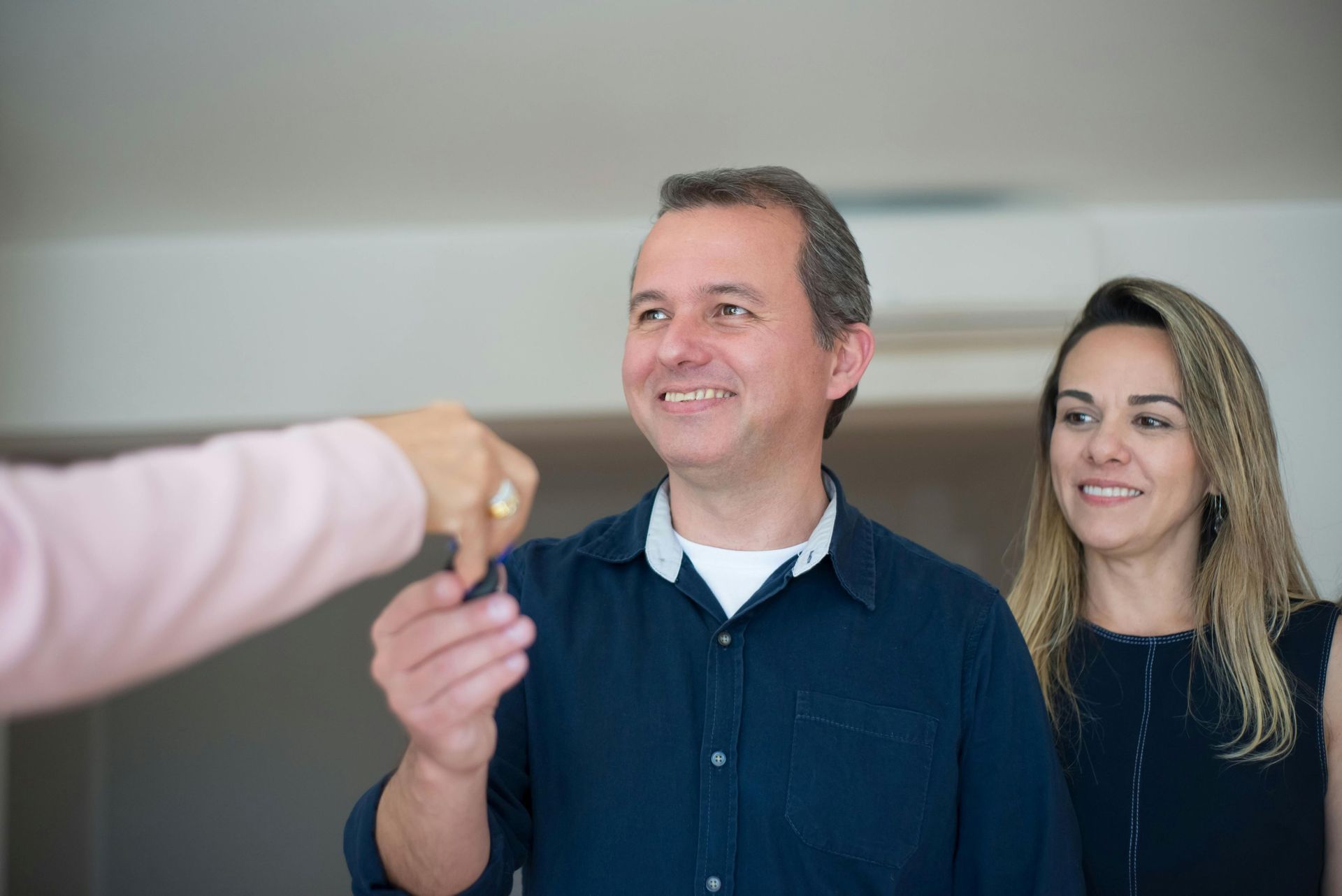 Man receiving keys from another person, smiling, with a woman beside him; indoors, neutral background.
