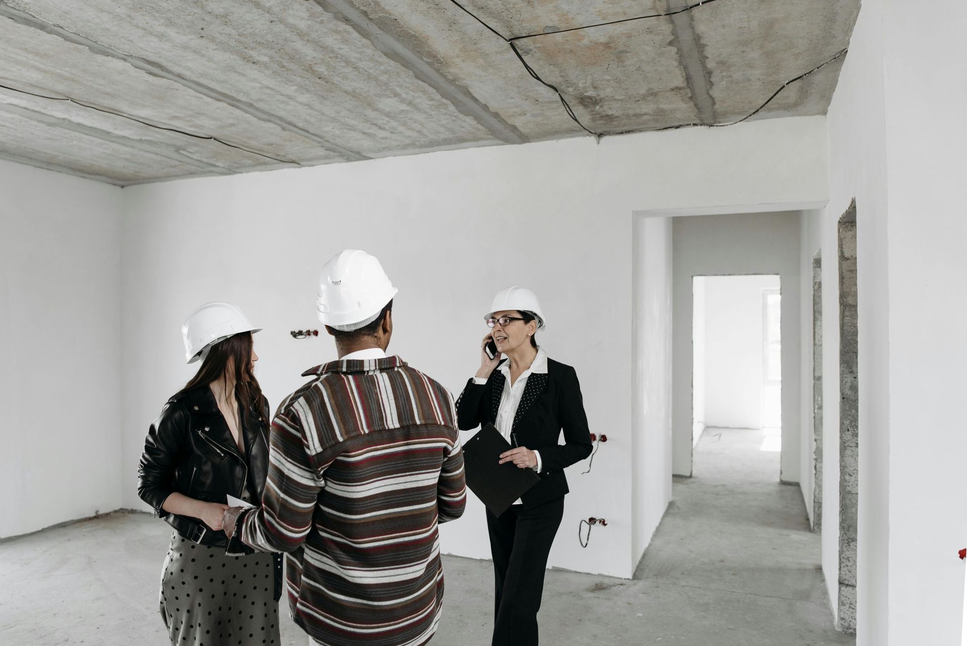 Three people wearing hard hats in an unfinished room. One on phone, others looking around.