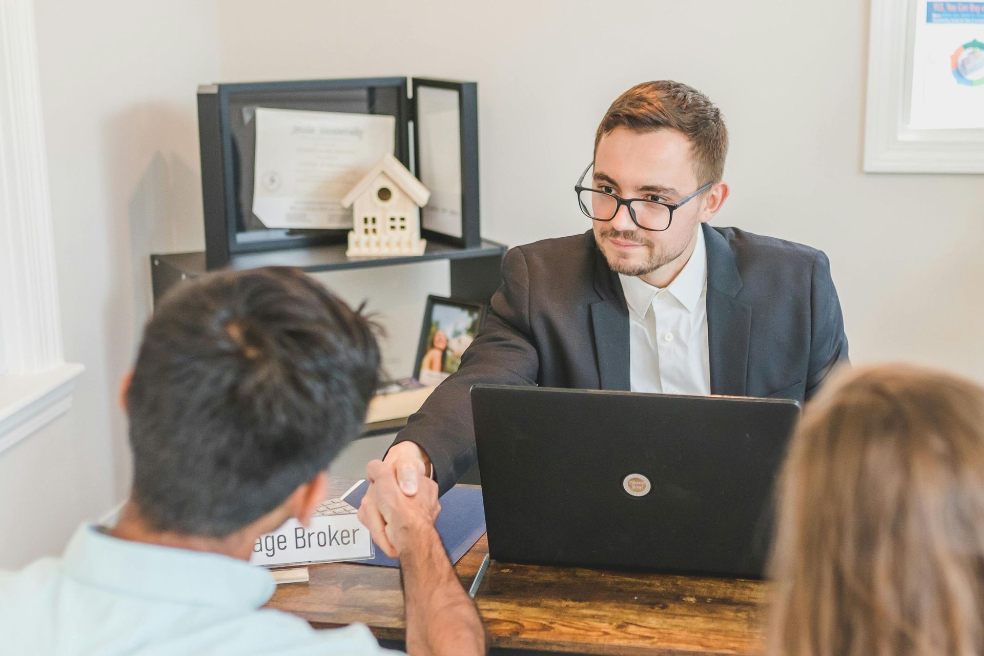Man in suit shakes hands with another man at a desk with a laptop and documents.