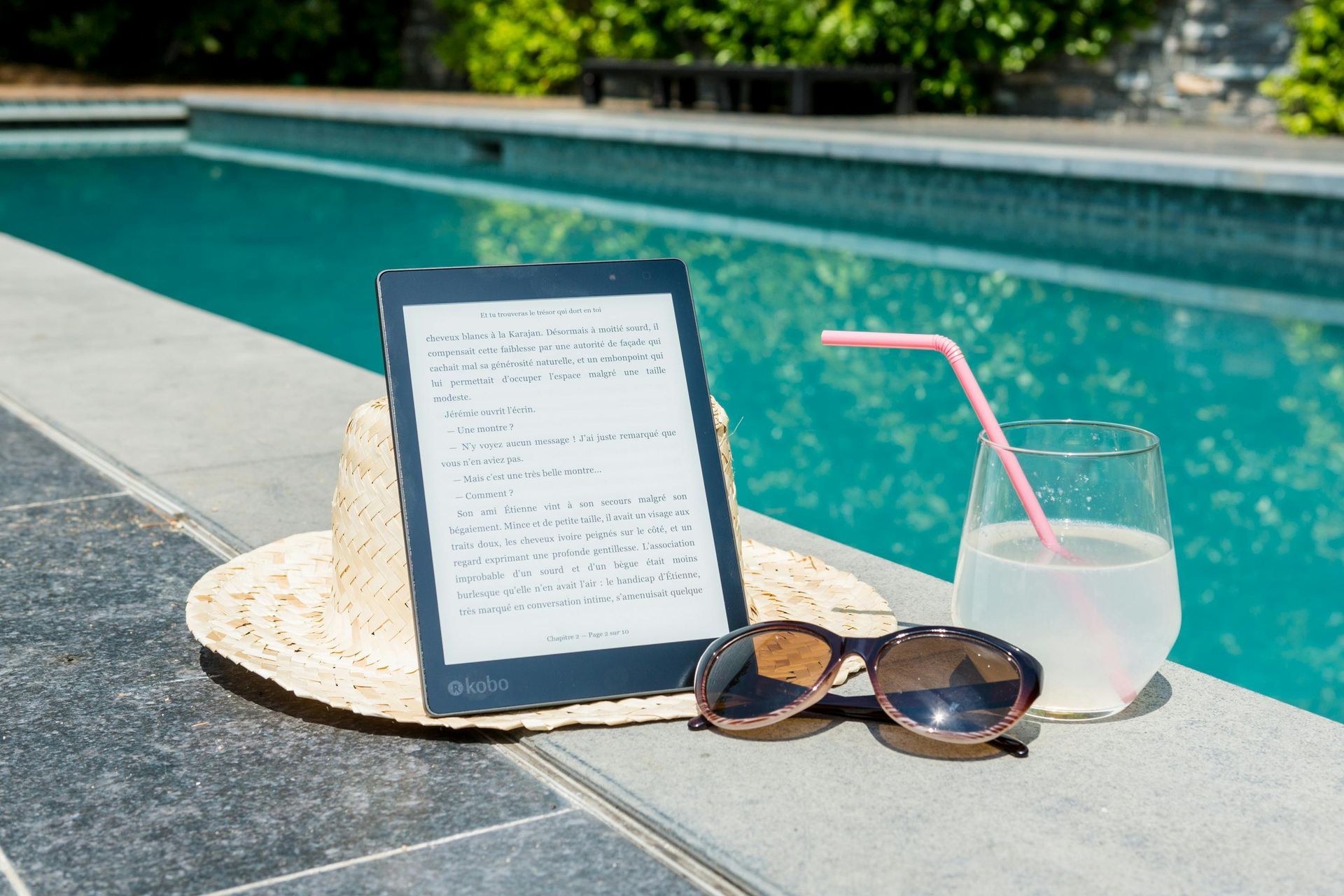 E-reader on straw hat next to sunglasses and a drink, poolside on a sunny day.