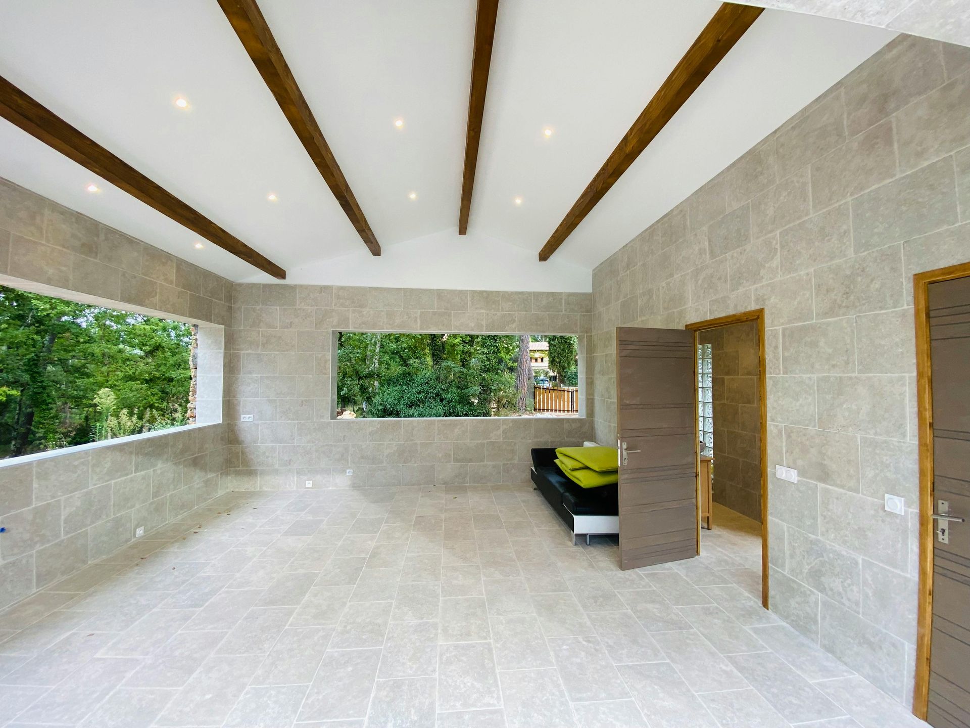 Empty room with tile floor, stone walls, wood beams, and large windows overlooking trees.