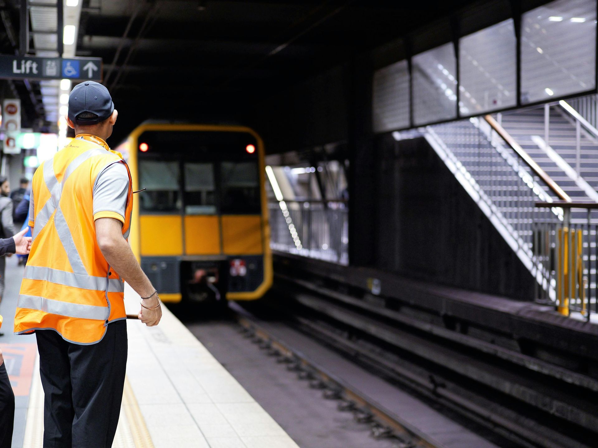 A person in a safety vest stands on a train platform as a yellow train approaches.