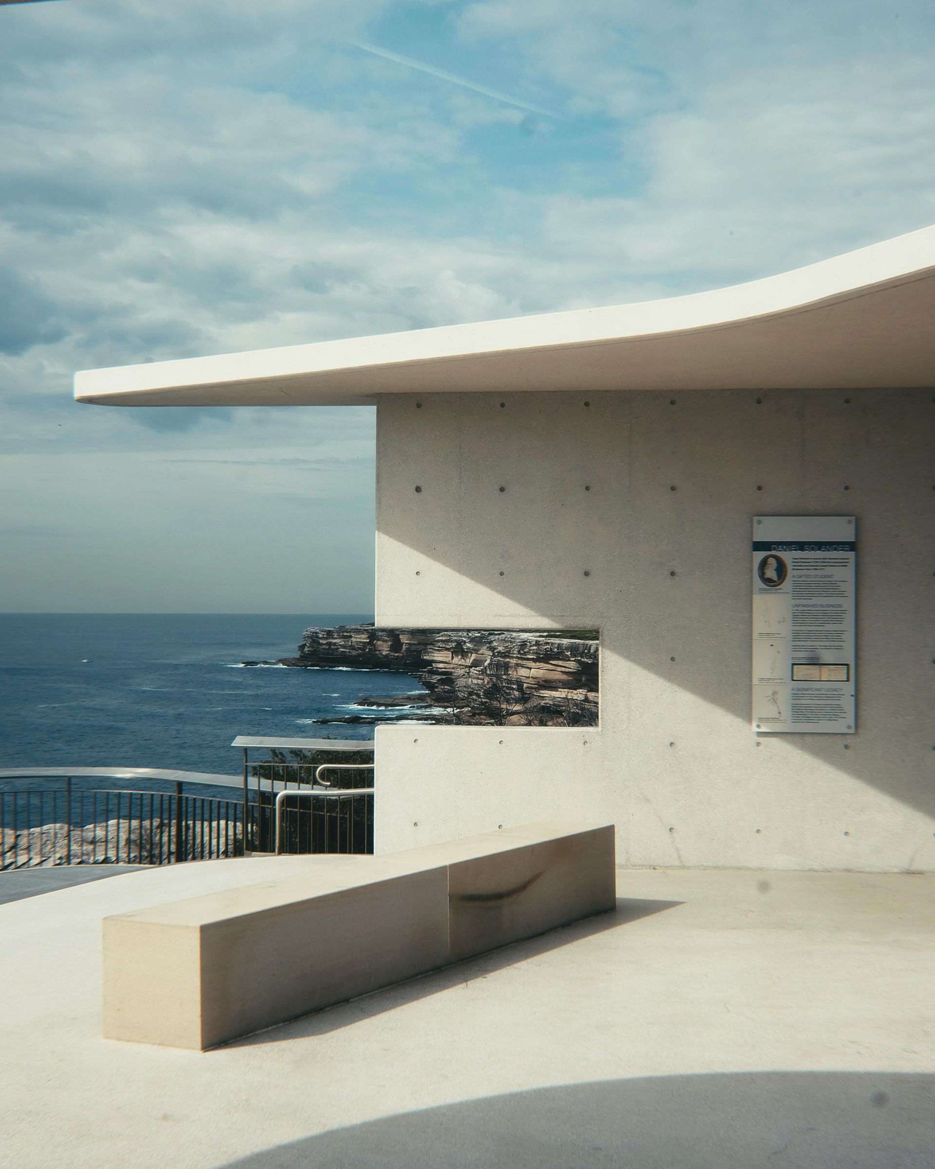 Bench and wall overlook ocean; signage, cityscape artwork, and curved roof. Sky is blue with clouds.