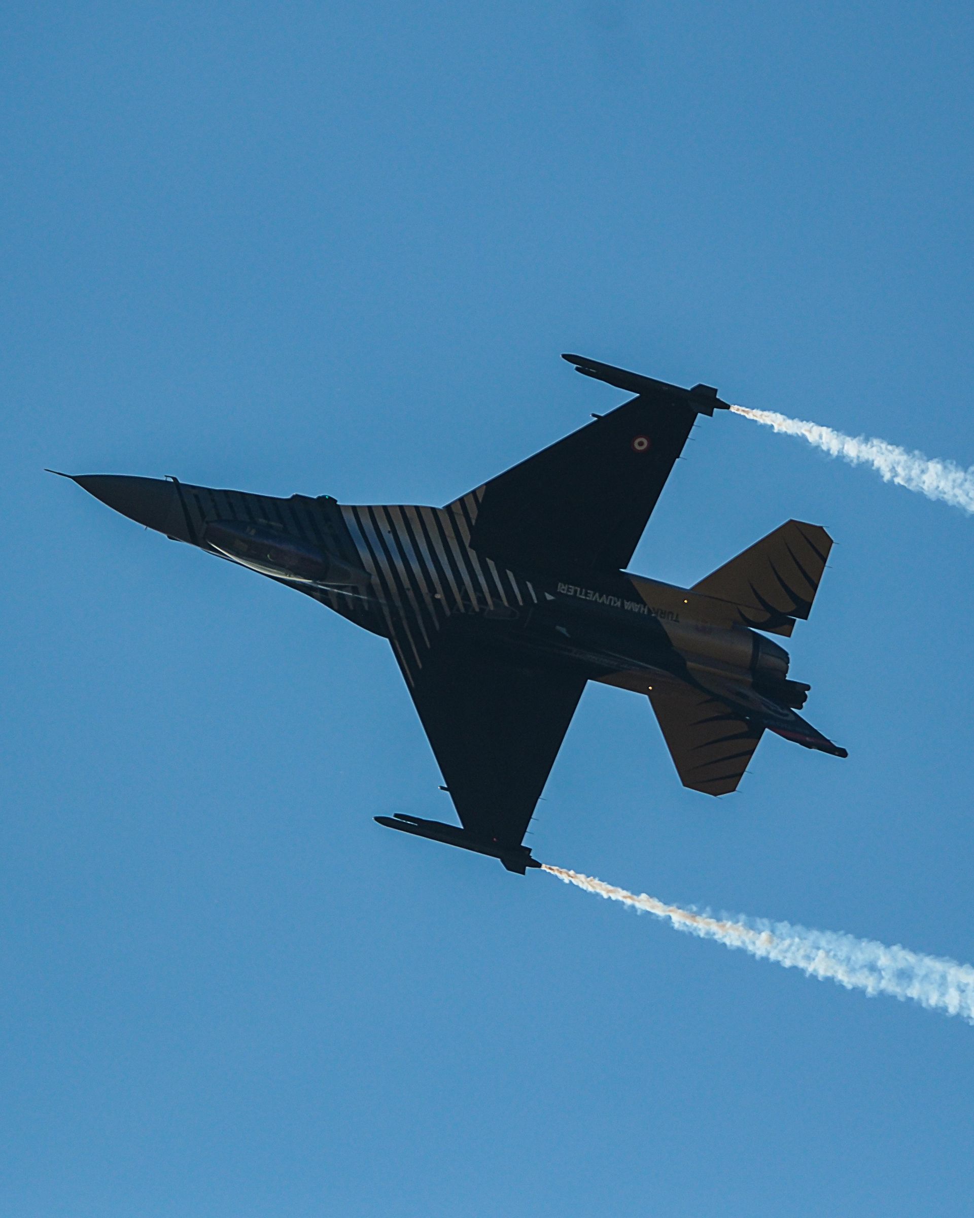 Black fighter jet in flight against a clear blue sky, leaving white vapor trails.