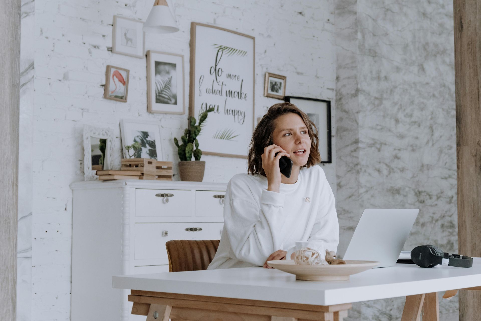 Woman talks on the phone, leaning on a white desk with laptop. White brick wall background with art.