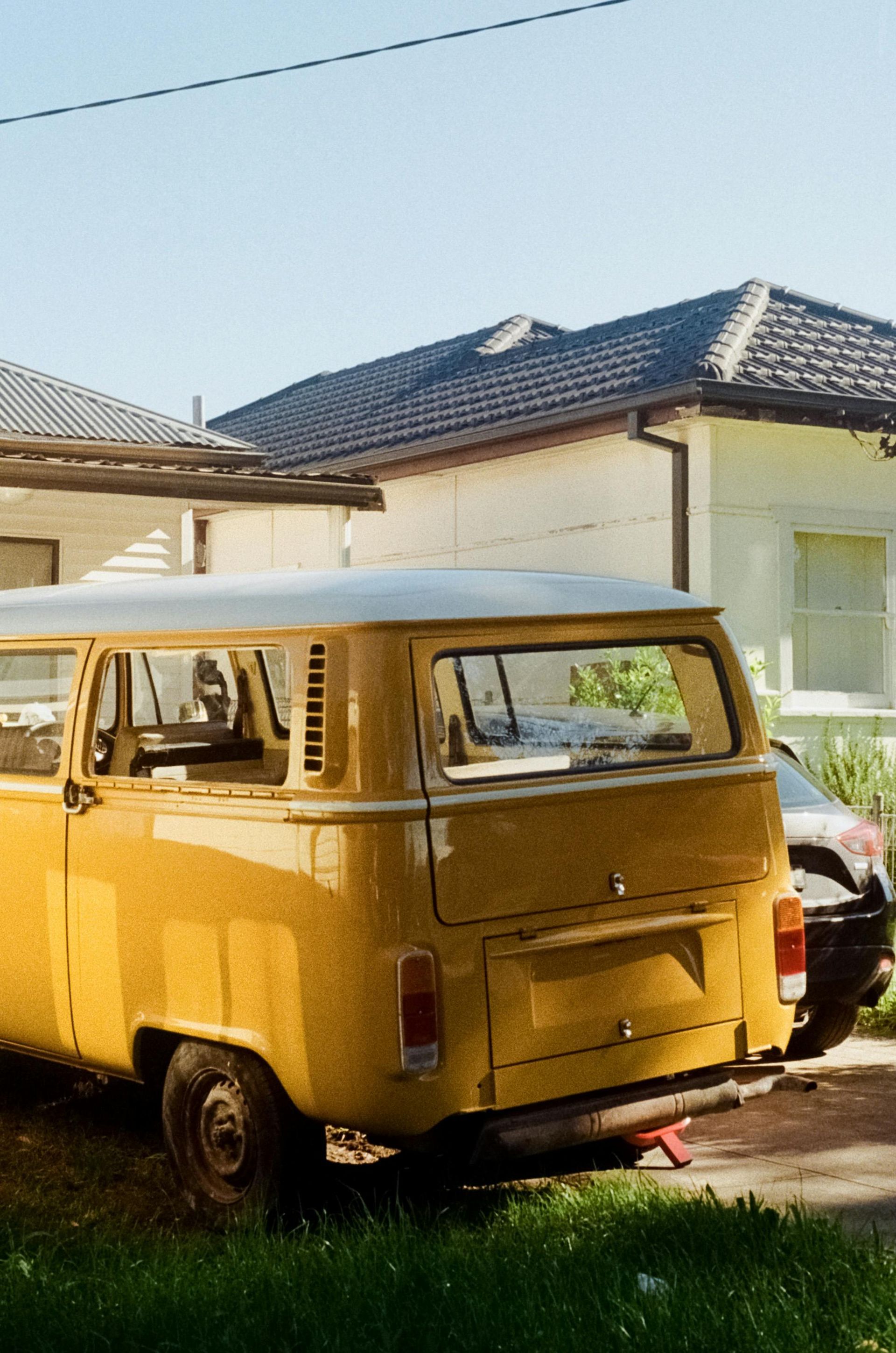 Yellow vintage Volkswagen van parked in front of a white house with a dark roof.