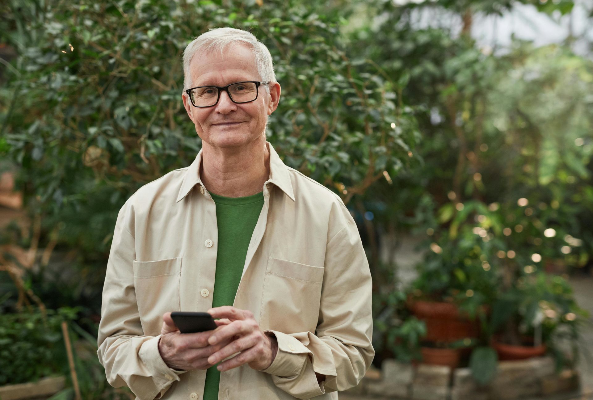 Man in glasses, holding phone, smiles in a greenhouse surrounded by lush green plants.