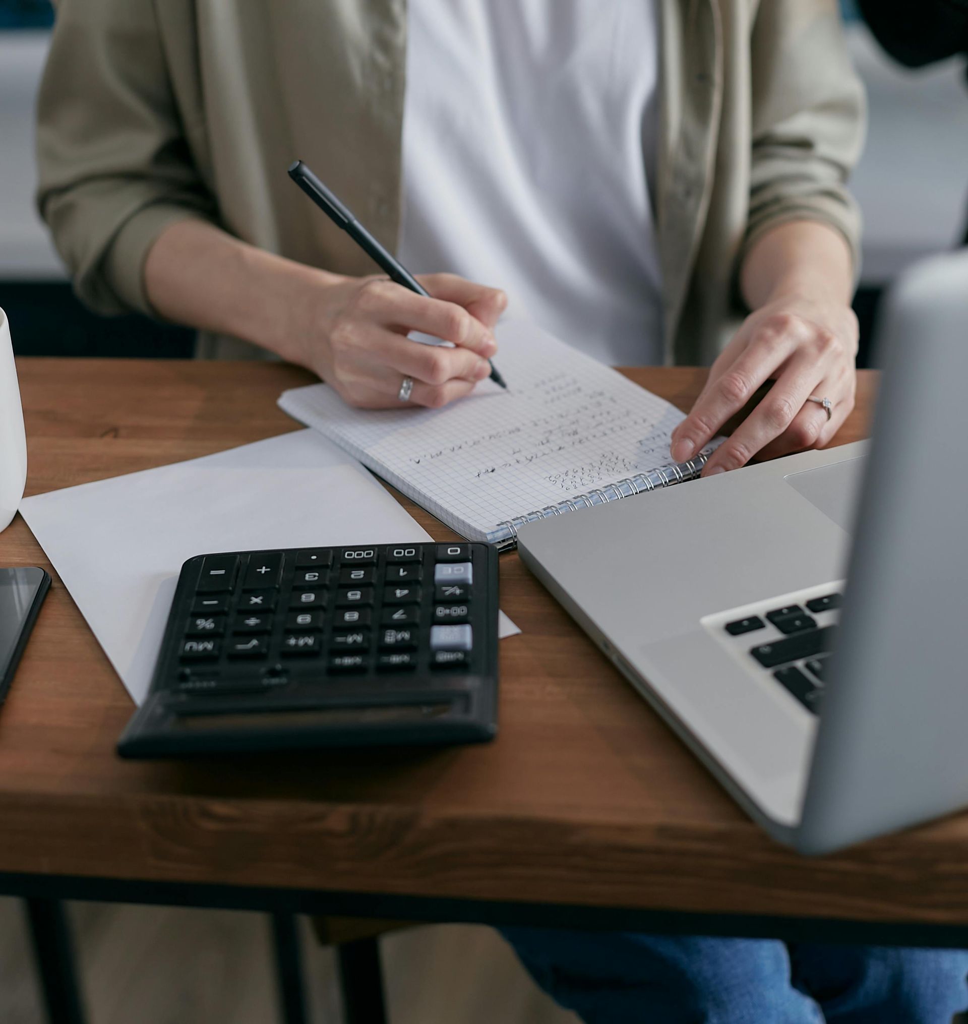 Person writing in a notebook, with a calculator and laptop on a desk.