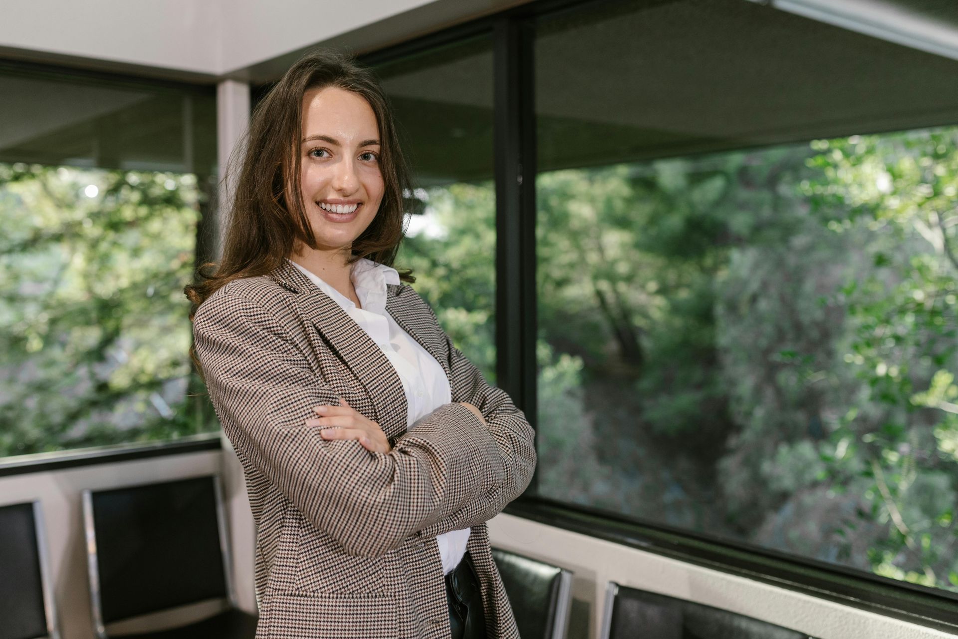 Woman in patterned blazer smiles, arms crossed, by a window overlooking trees.