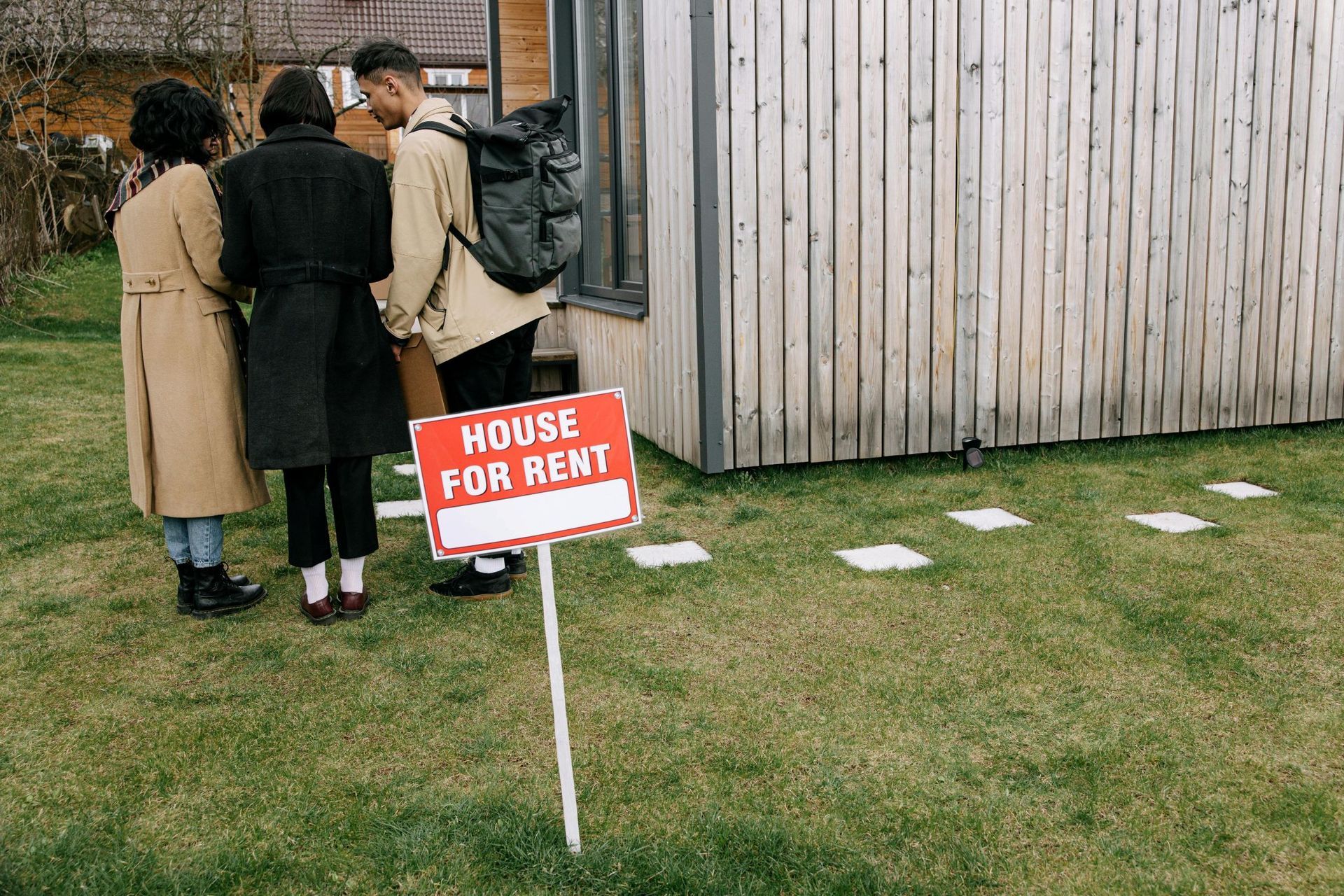 Three people looking at a house for rent sign in front of a small wooden house.