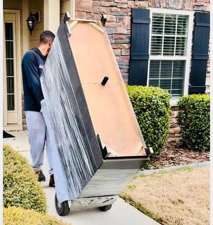 Person carries a large cabinet on a dolly near a house with brick siding and a front door.