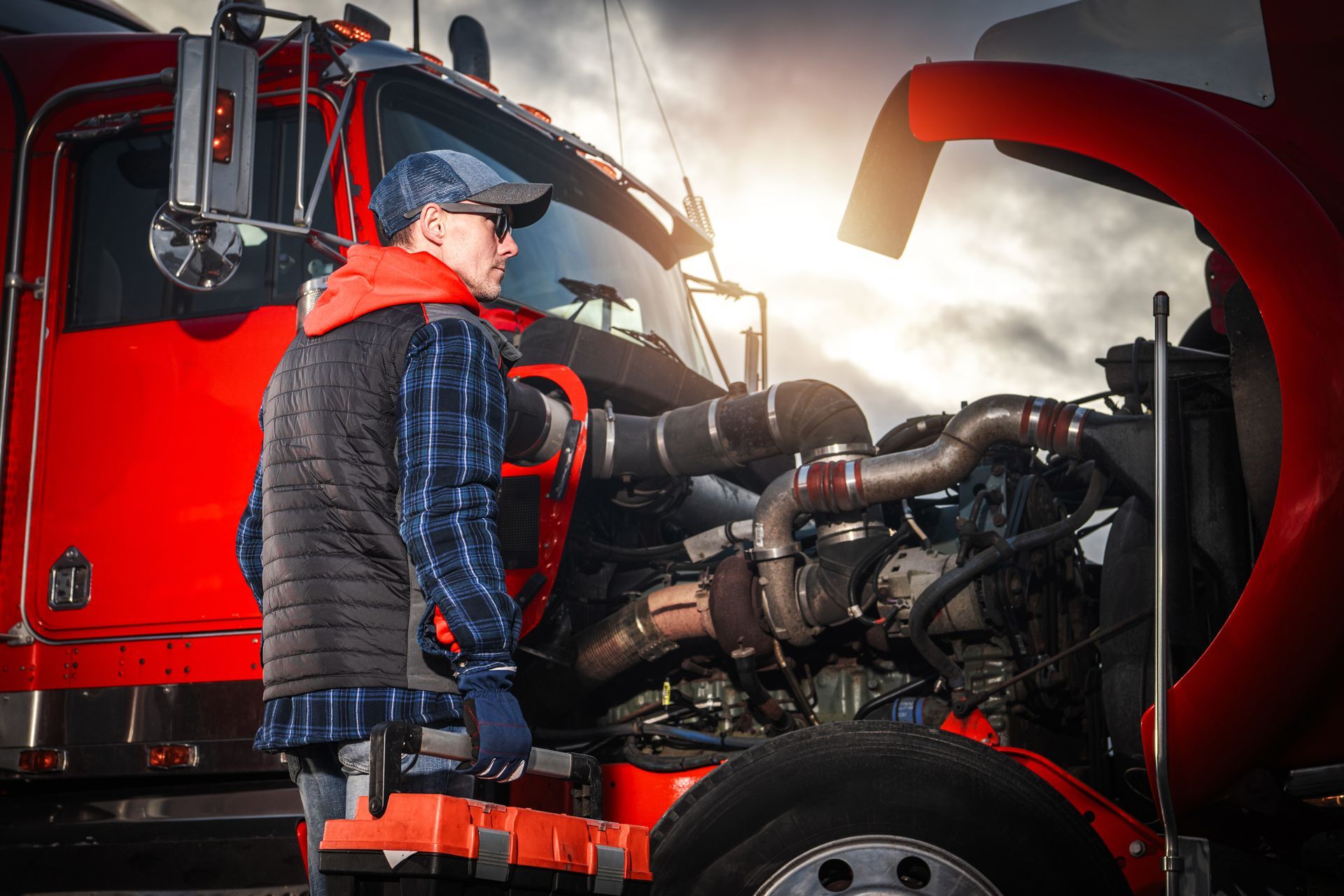 Mechanic inspecting a truck engine beside a red semi-truck in a garage