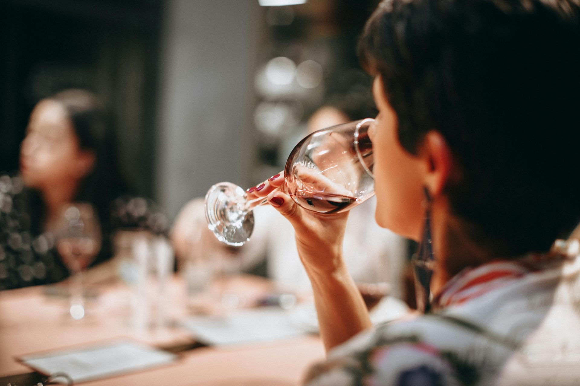 Woman sipping red wine at a table; other people and glasses are visible in a blurred background.