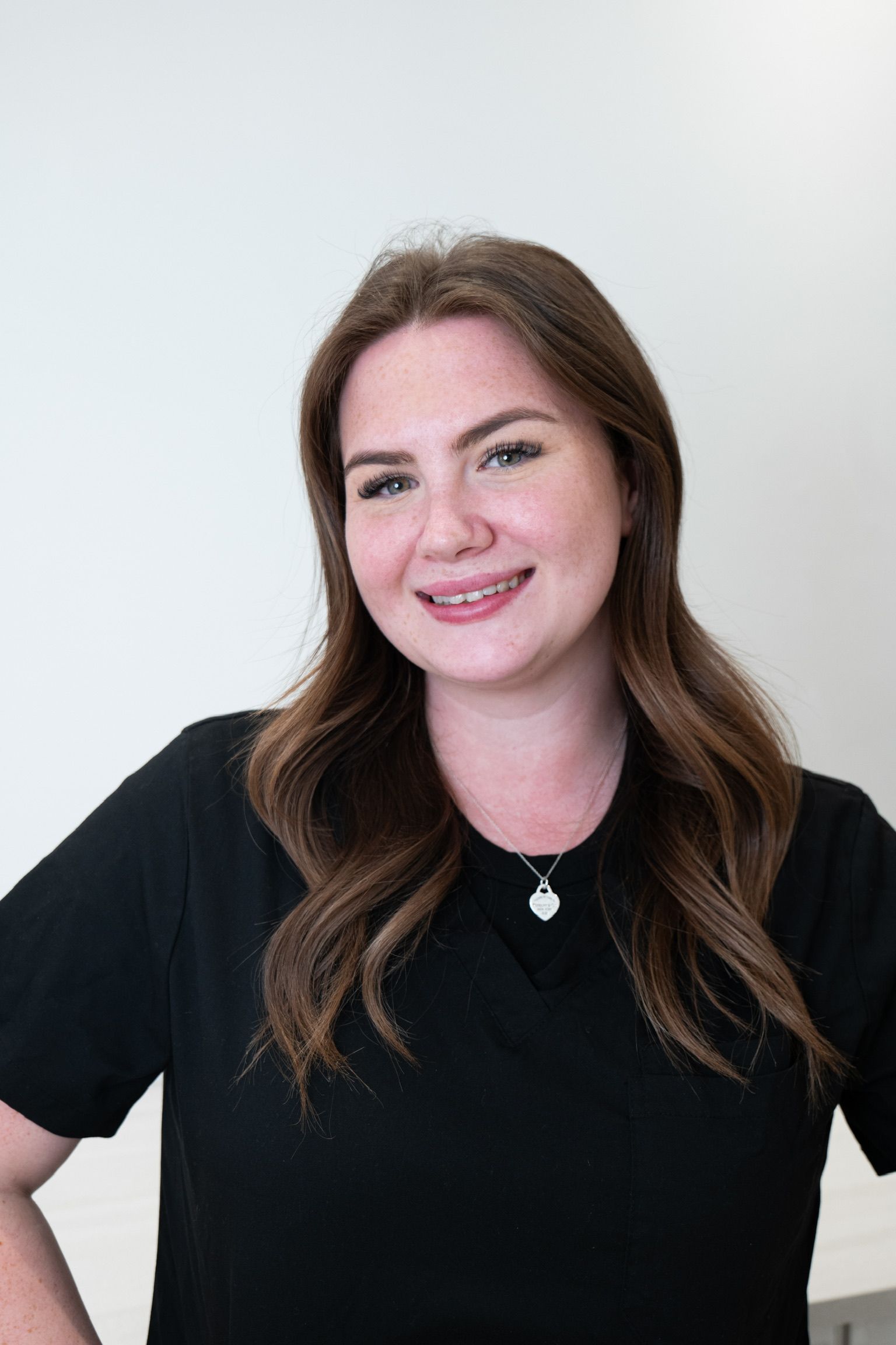A woman wearing a black shirt and a necklace is standing in front of a white wall.