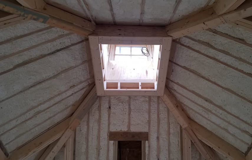 An interior view of a wooden attic frame with foam insulation between beams and a square skylight open to the sky.