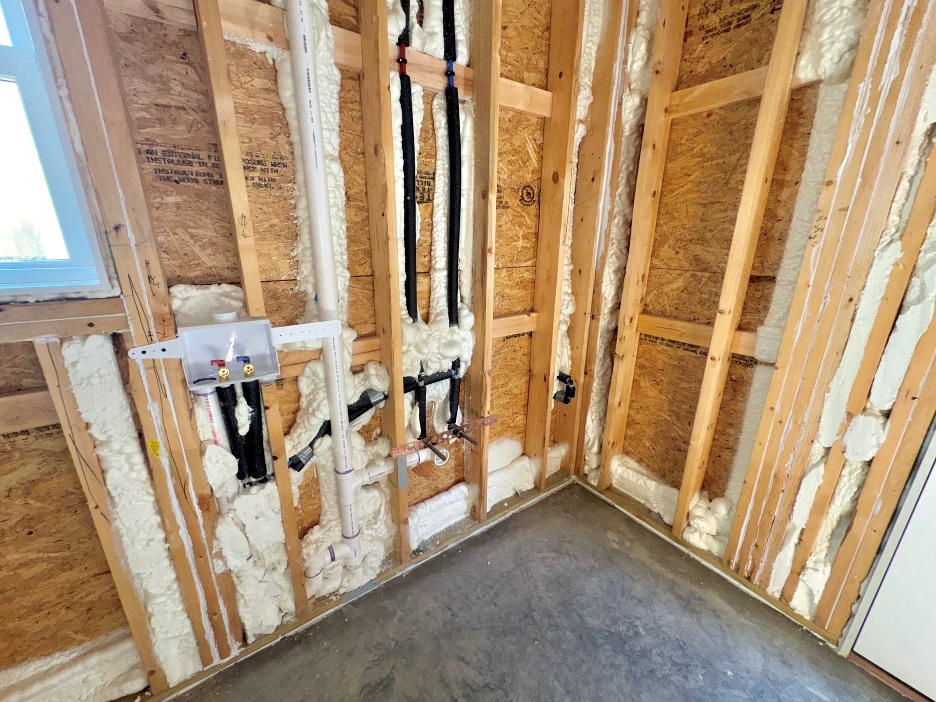 Wall framing and spray foam insulation in an unfinished room with exposed plumbing for a laundry hookup and pipes.