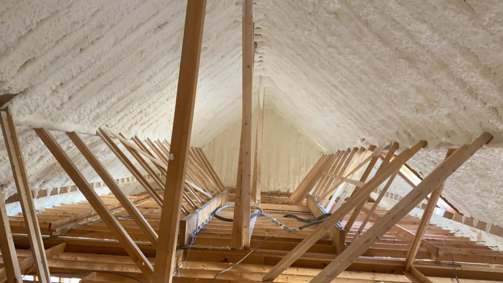 An interior view of a wooden attic frame with foam insulation between beams and a square skylight open to the sky.