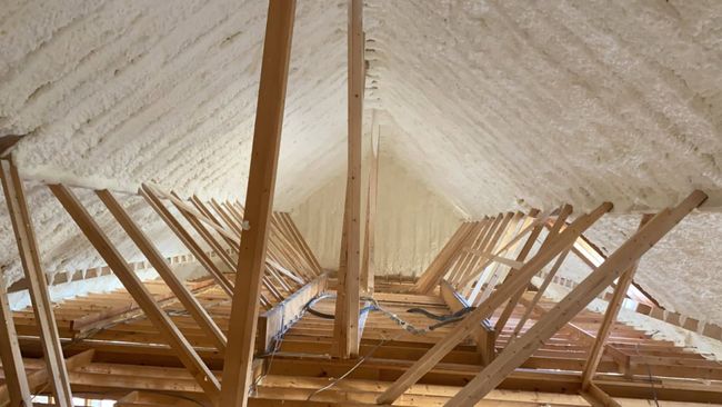 An interior view of a wooden attic frame with foam insulation between beams and a square skylight open to the sky.