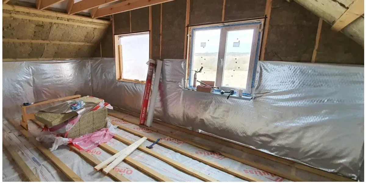 Attic under renovation with fiberglass insulation, silver vapor barrier, and timber floor joists.