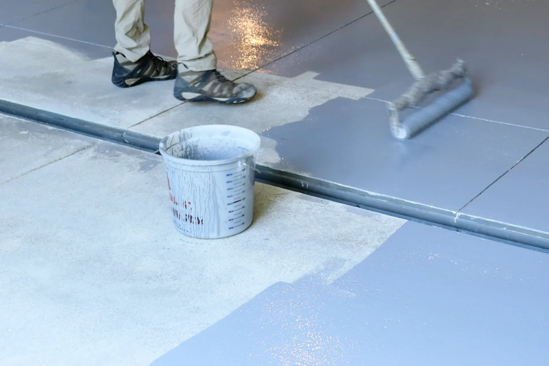 A person uses a paint roller to apply grey epoxy coating to a concrete floor, with a bucket sitting nearby.