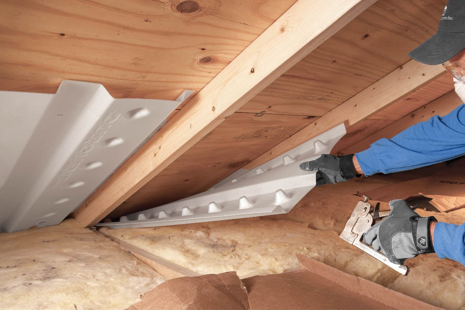 A worker installs white plastic rafter vents between wooden roof joists in an attic to ensure proper insulation airflow.