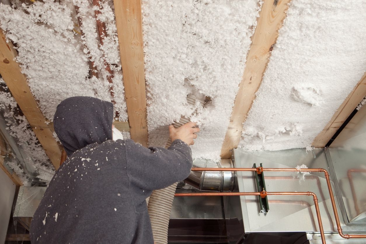 A person wearing a hooded sweatshirt sprays white insulation foam between wooden ceiling joists near copper pipes.