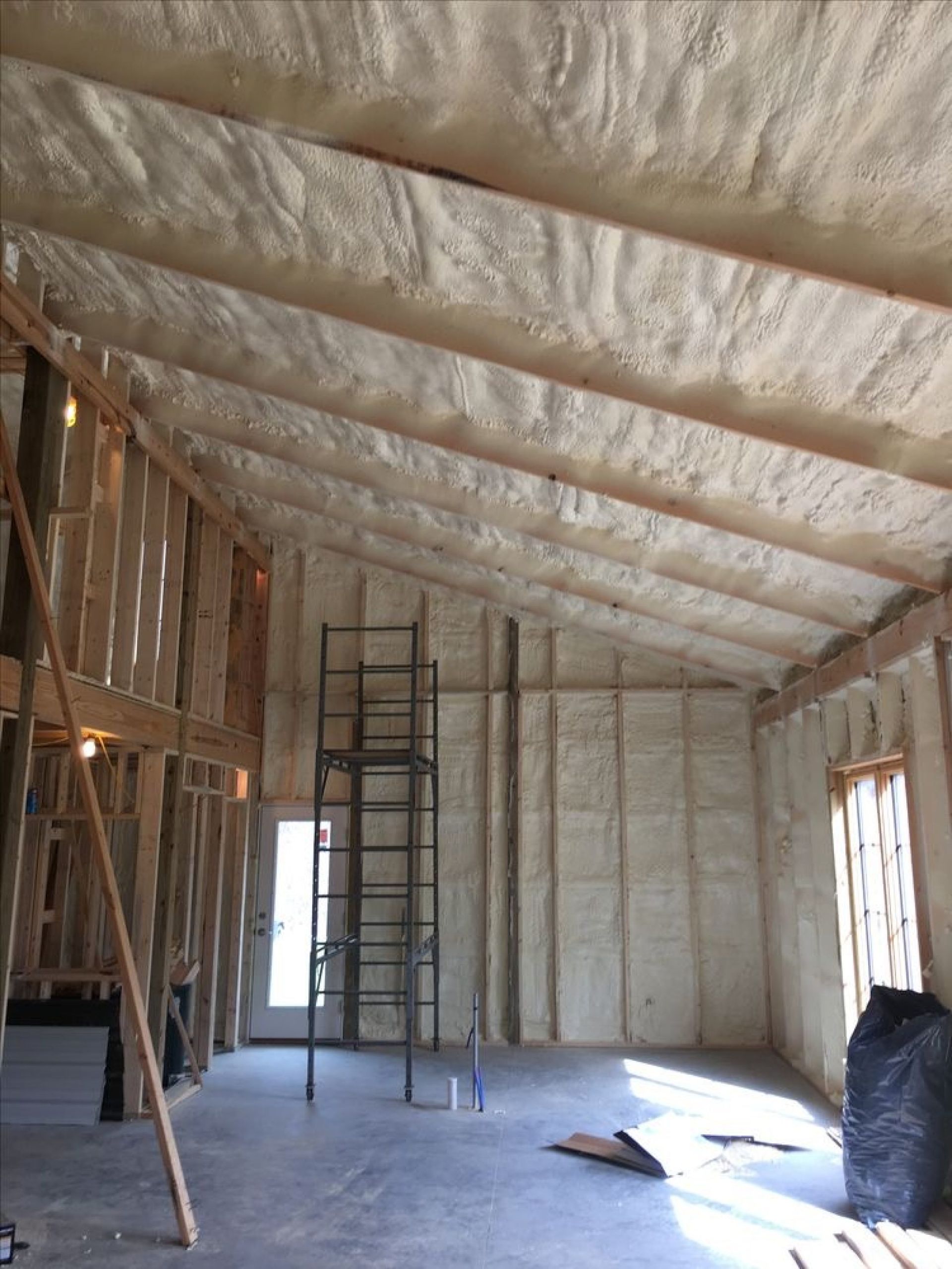 Interior of a room under construction with white spray foam insulation on the walls and vaulted ceiling, and a ladder.