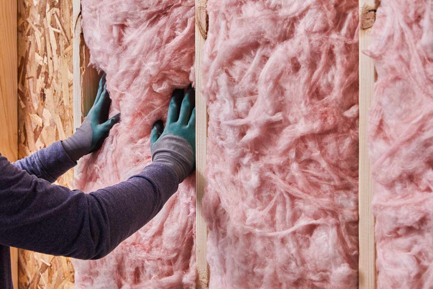 A person wearing gloves presses pink fiberglass batt insulation between wooden wall studs.