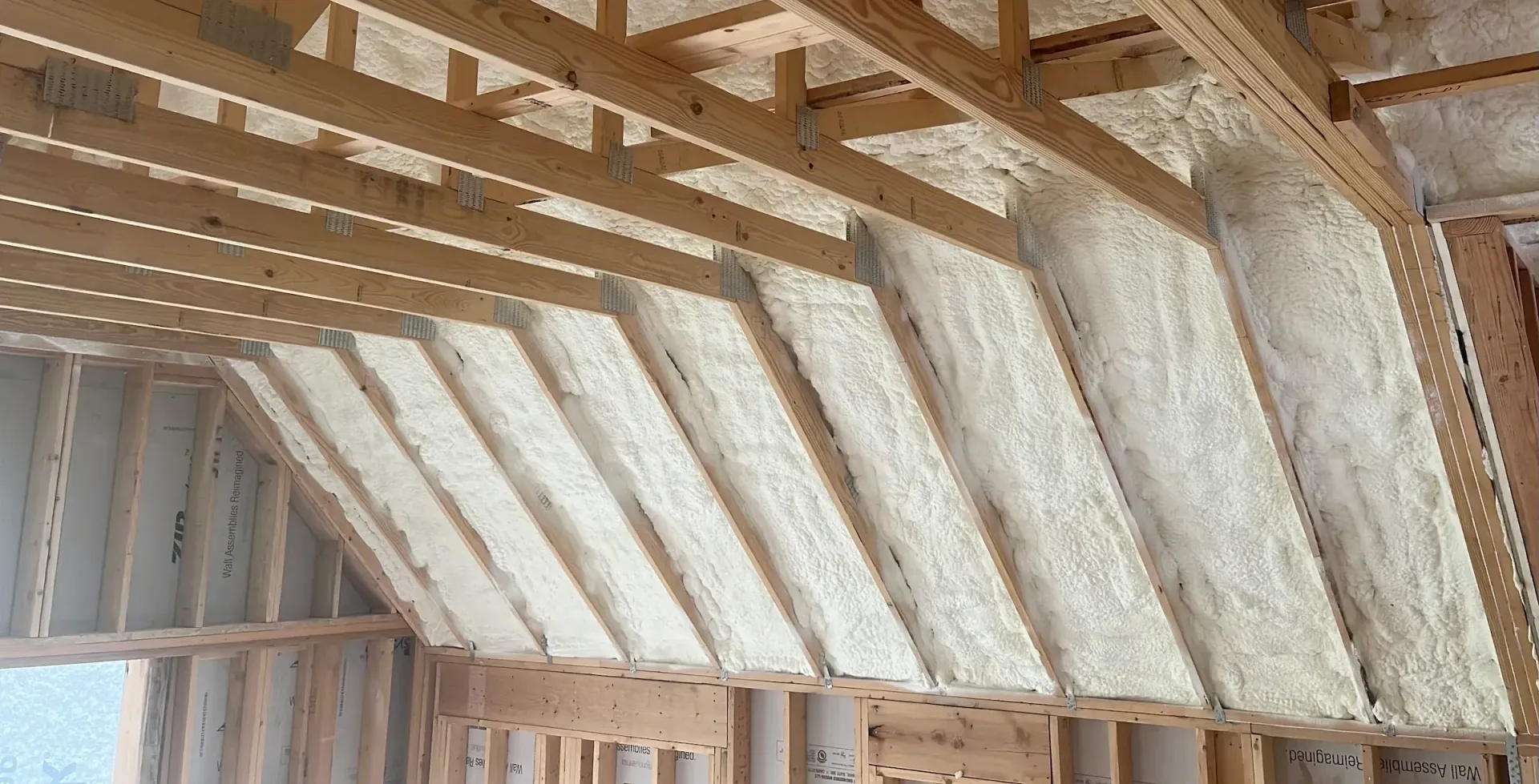An interior view of a wooden attic frame with foam insulation between beams and a square skylight open to the sky.