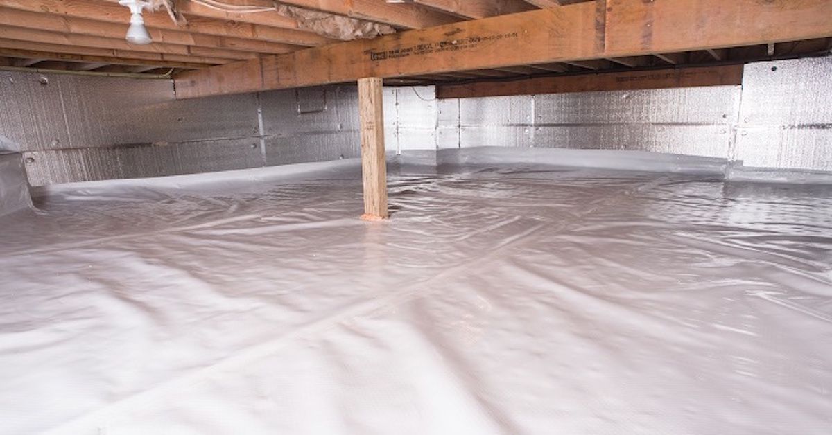 A person in a hard hat standing on a ladder and spraying white foam insulation onto an unfinished interior wall.