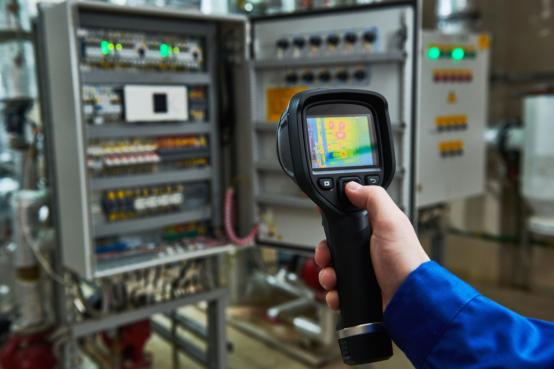 A person is holding a thermal camera in front of a control panel.