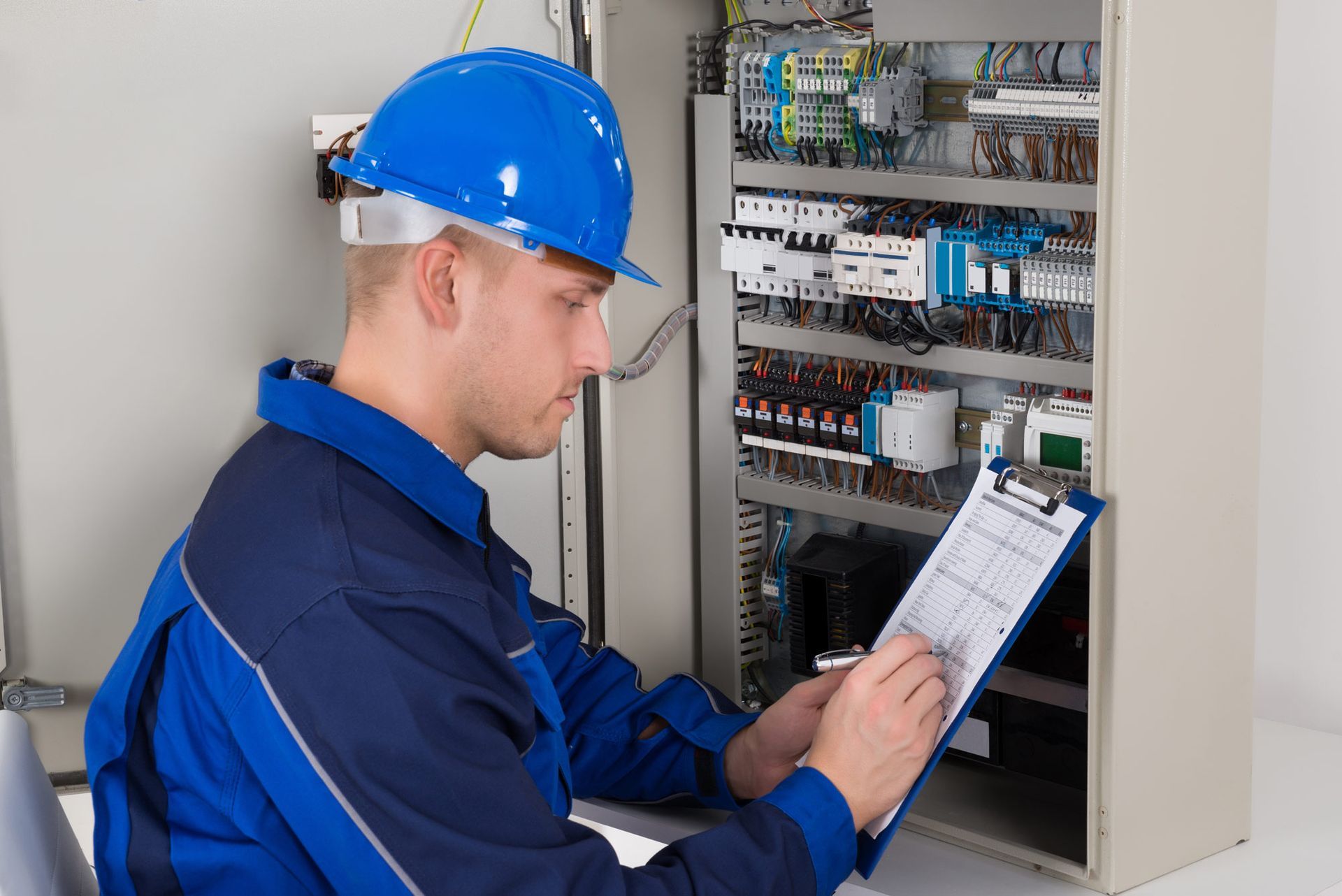 A man wearing a hard hat is looking at a clipboard in front of an electrical cabinet.