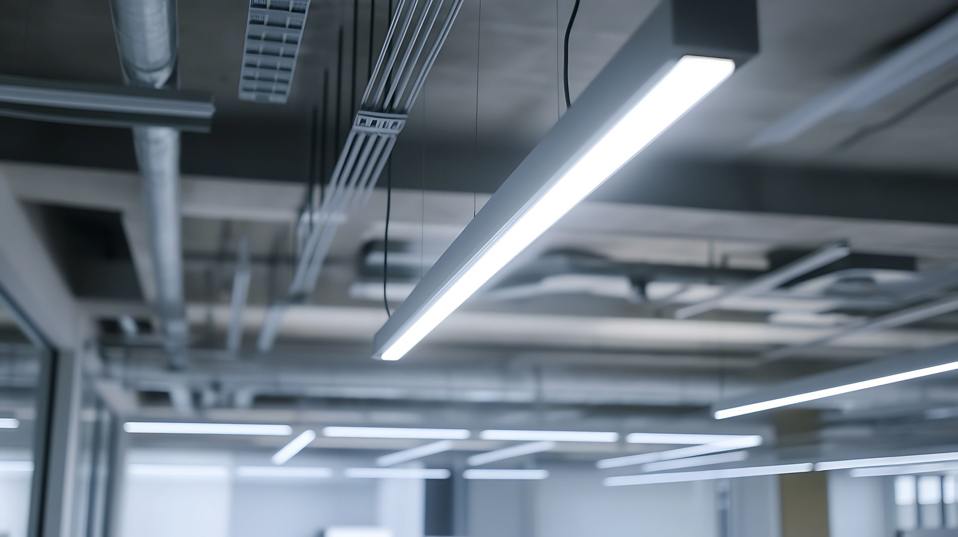 Bright, linear LED light fixture on a concrete ceiling with exposed pipes and conduits.