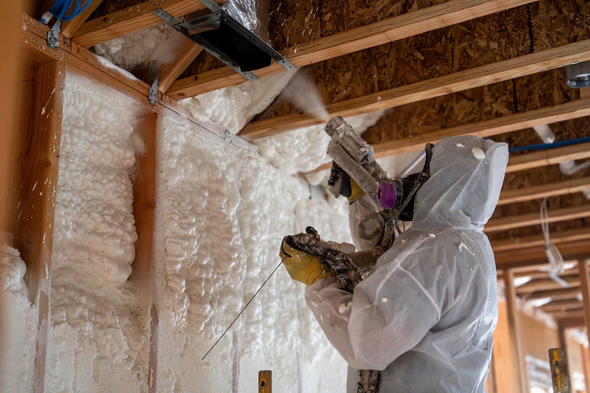 Worker in protective gear spraying foam insulation on wooden walls inside a house frame.