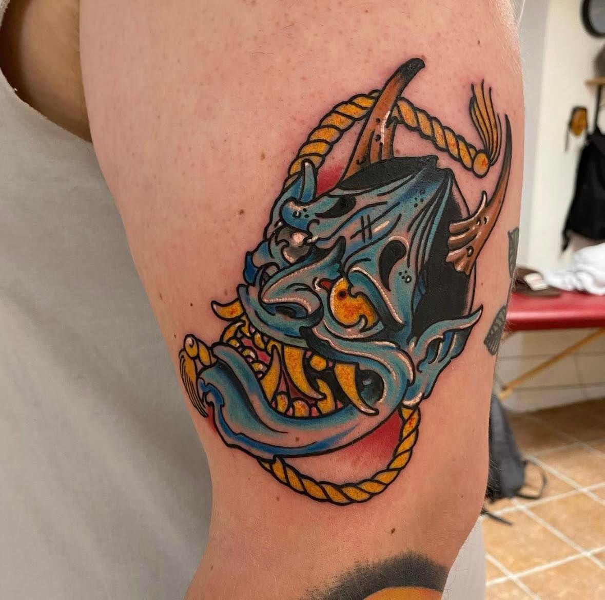 Fargerik japansk-inspirert tatovering av demonmaske med blå, gul og rød fargepalett – på overarm