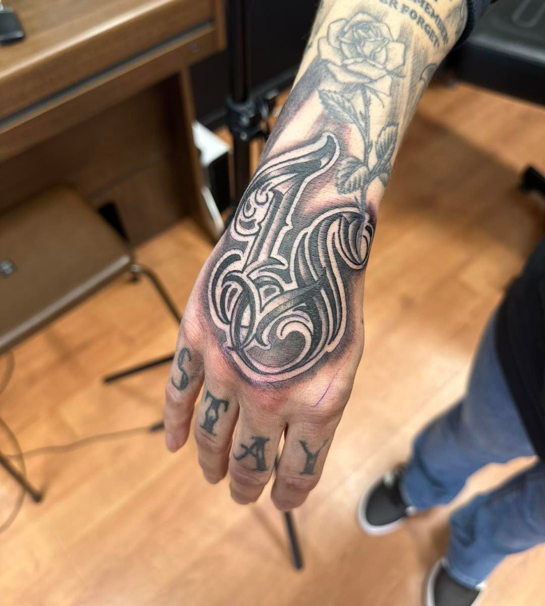 Detaljert håndtatovering med gotisk bokstav L, rose og teksten STAY på fingrene