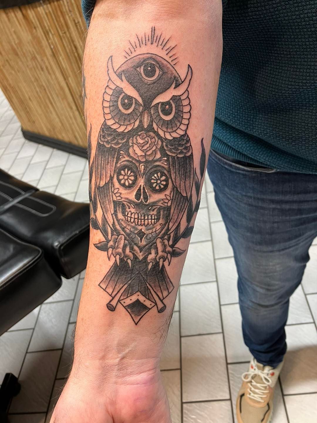 Sort og grå tatovering av mystisk ugle med tredje øye, sugar skull, roser og korslagte objekter – på underarm