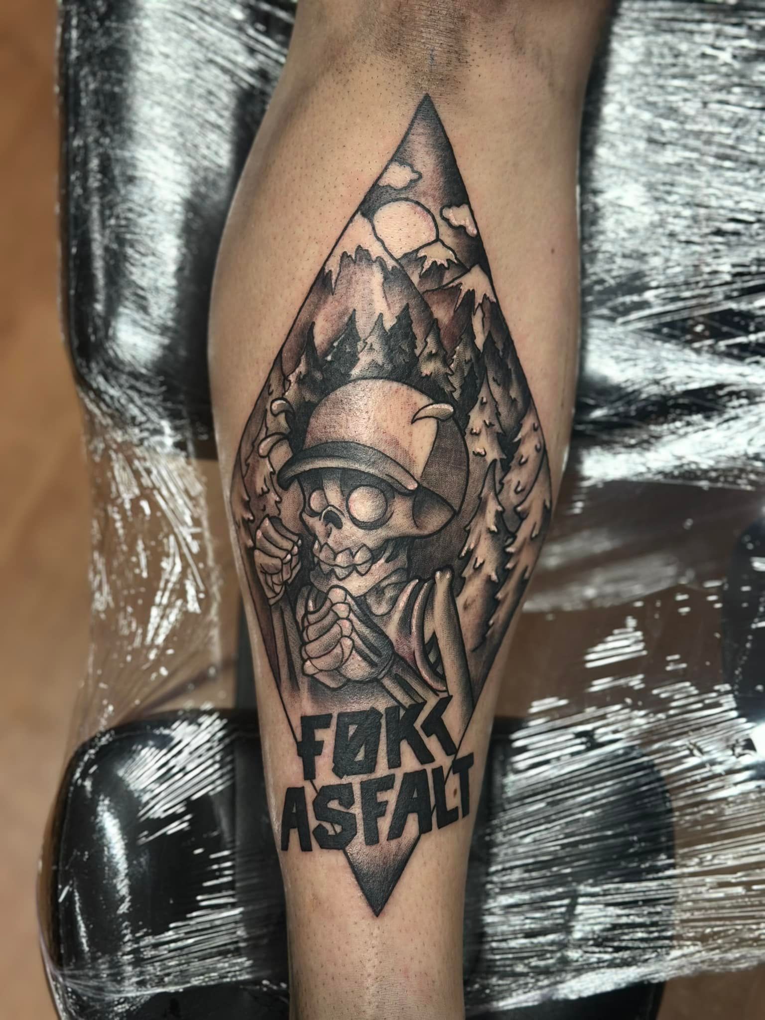 Blackwork tatovering av skjelett på sykkel i skoglandskap med teksten FØKK ASFALT – på legg