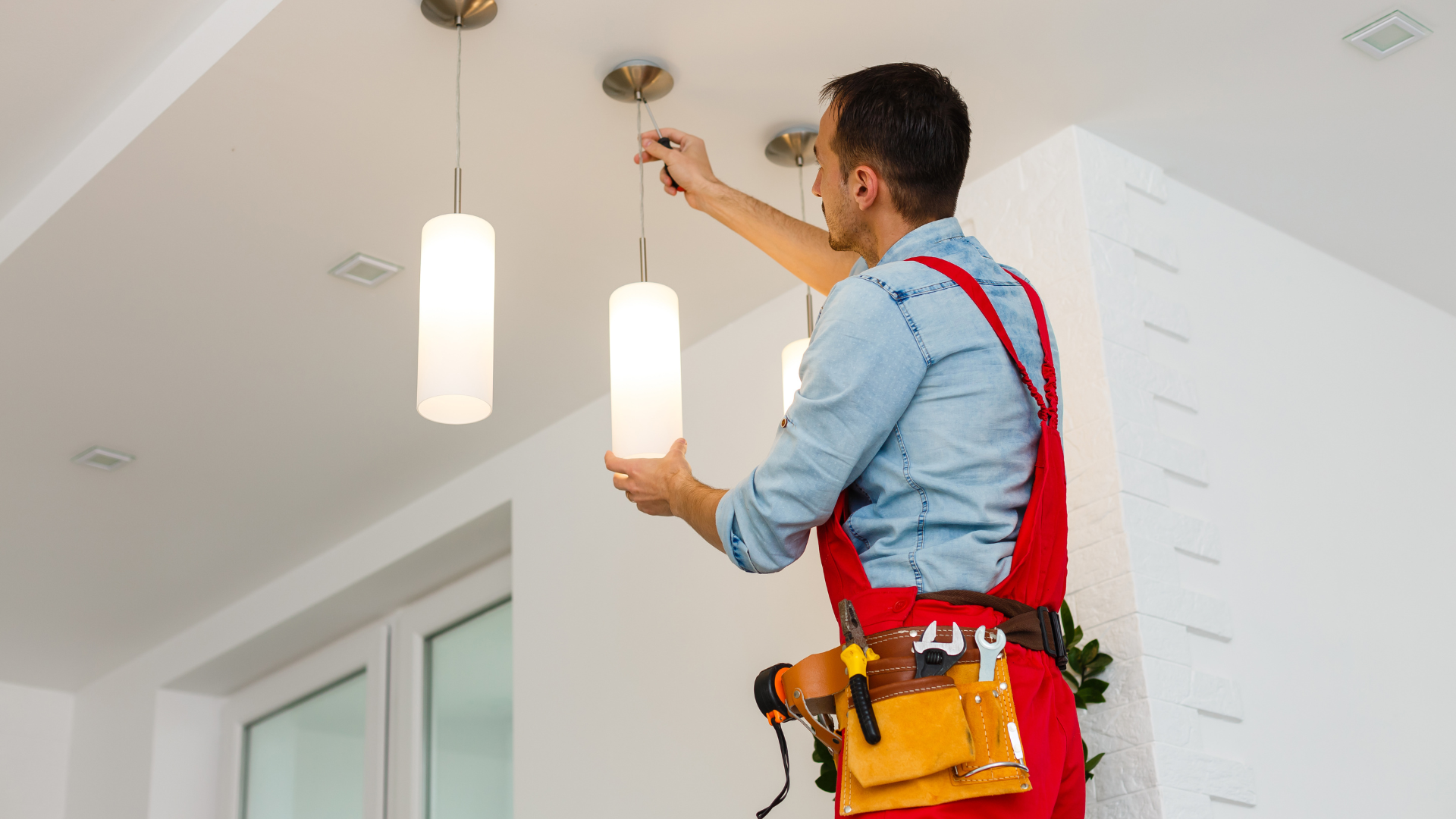 An electrician in red overalls is installing a pendant light fixture on a white ceiling.
