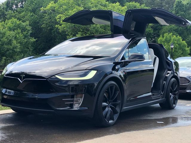 Black Tesla Model X with gullwing doors open, parked outdoors near greenery.