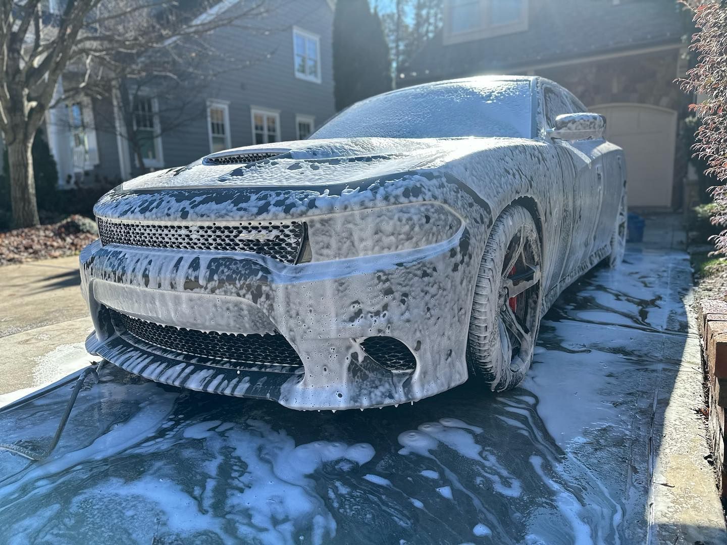 A black Dodge Charger covered in foamy soap suds sits on a driveway, ready for a wash.