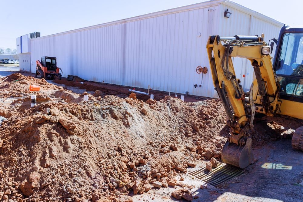 Yellow excavator beside a white industrial building, digging a large dirt trench at a construction site
