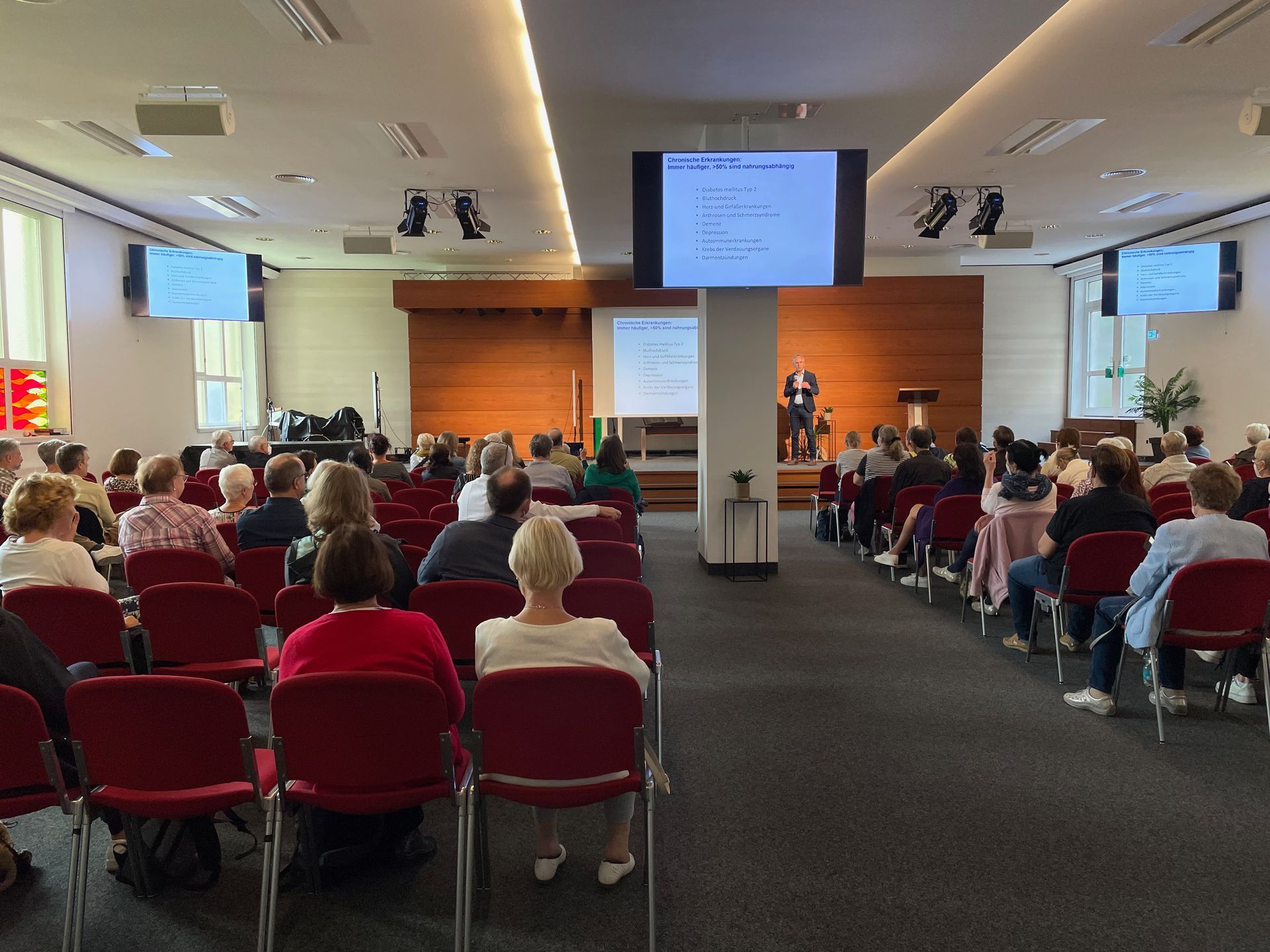 Ein großer Raum voller Menschen, die auf roten Stühlen sitzen und einer Präsentation zuschauen.