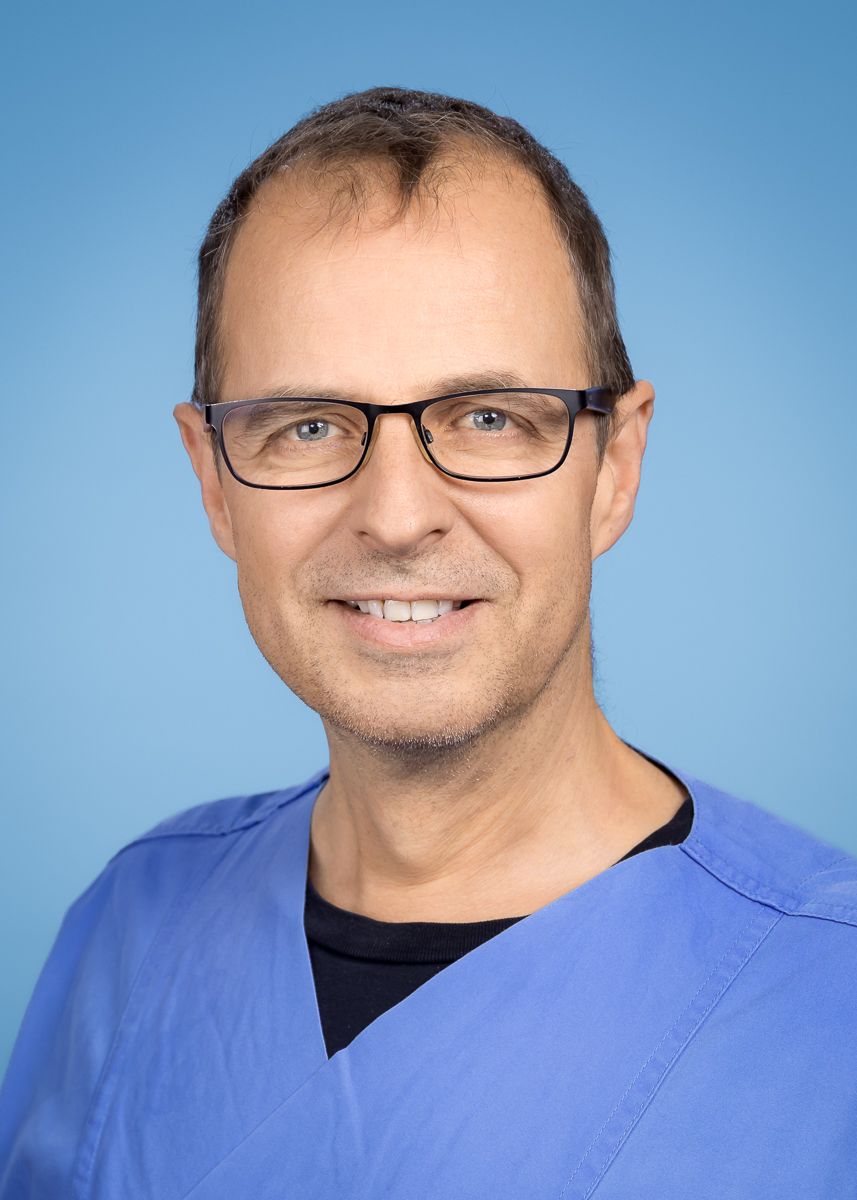 Ein Mann mit Brille und blauem OP-Kittel lächelt in die Kamera.